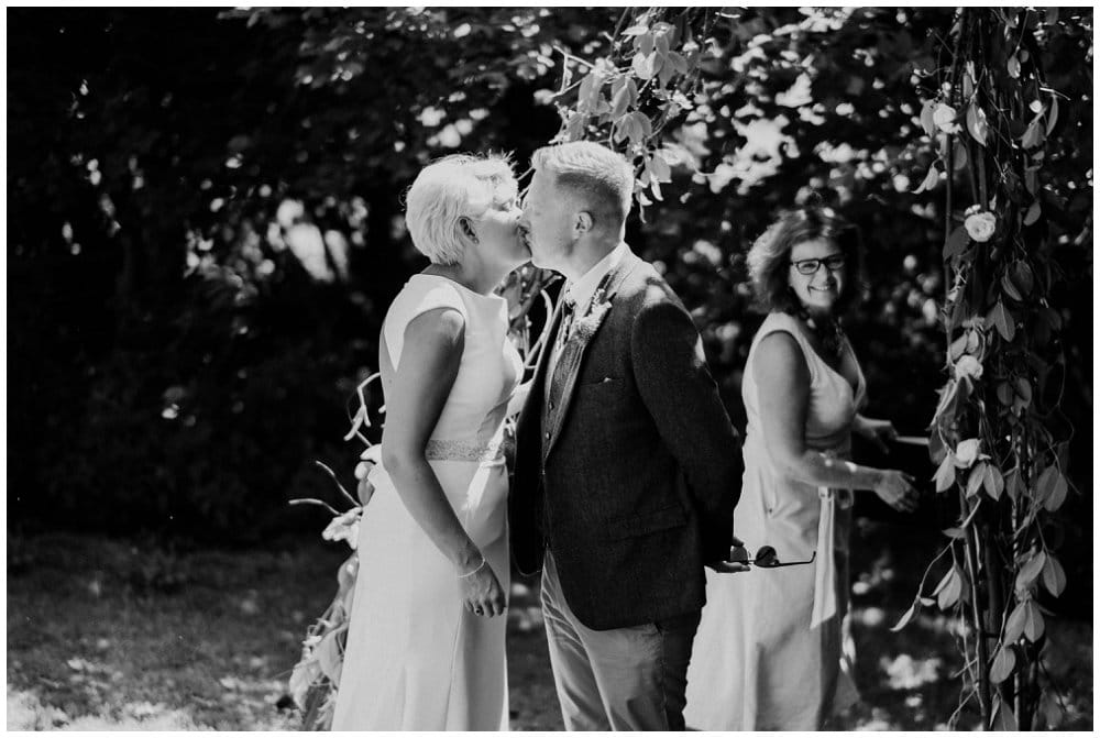 Celebrant for your wedding? black and white image of bride and groom kissing with Fan the celebrant looking on 