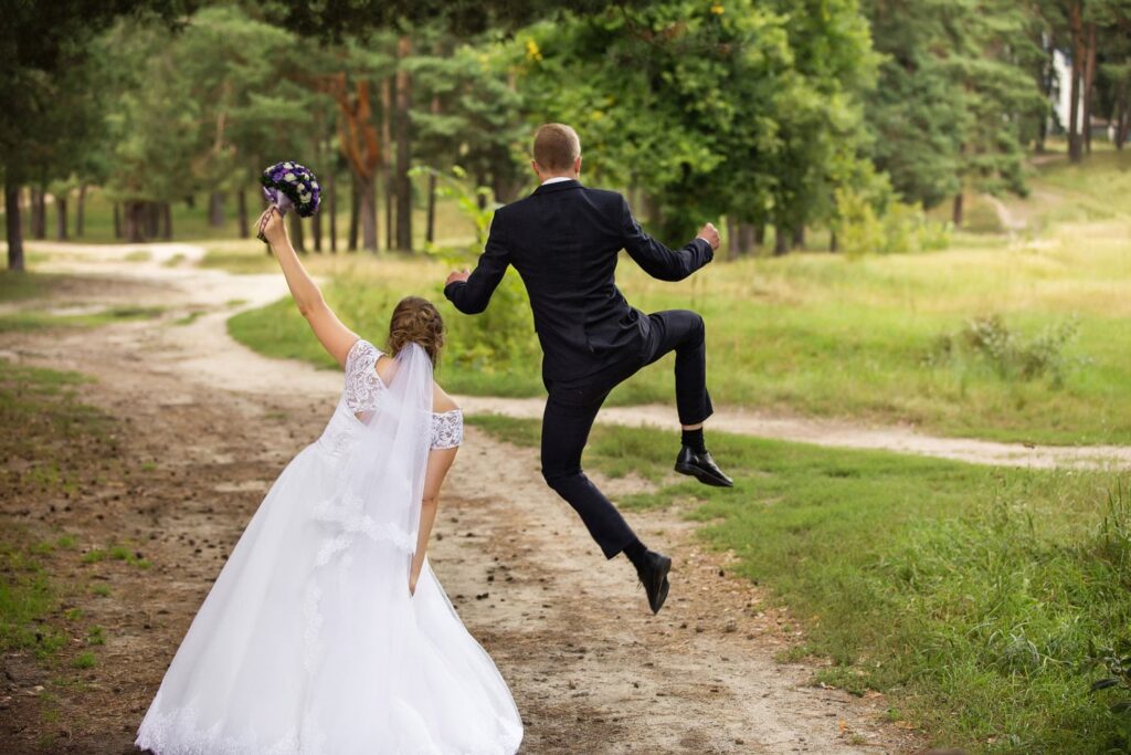 successful wedding planning a bride and groom walk away from camera up a leafy lane, the groom is doing a heel kick jump