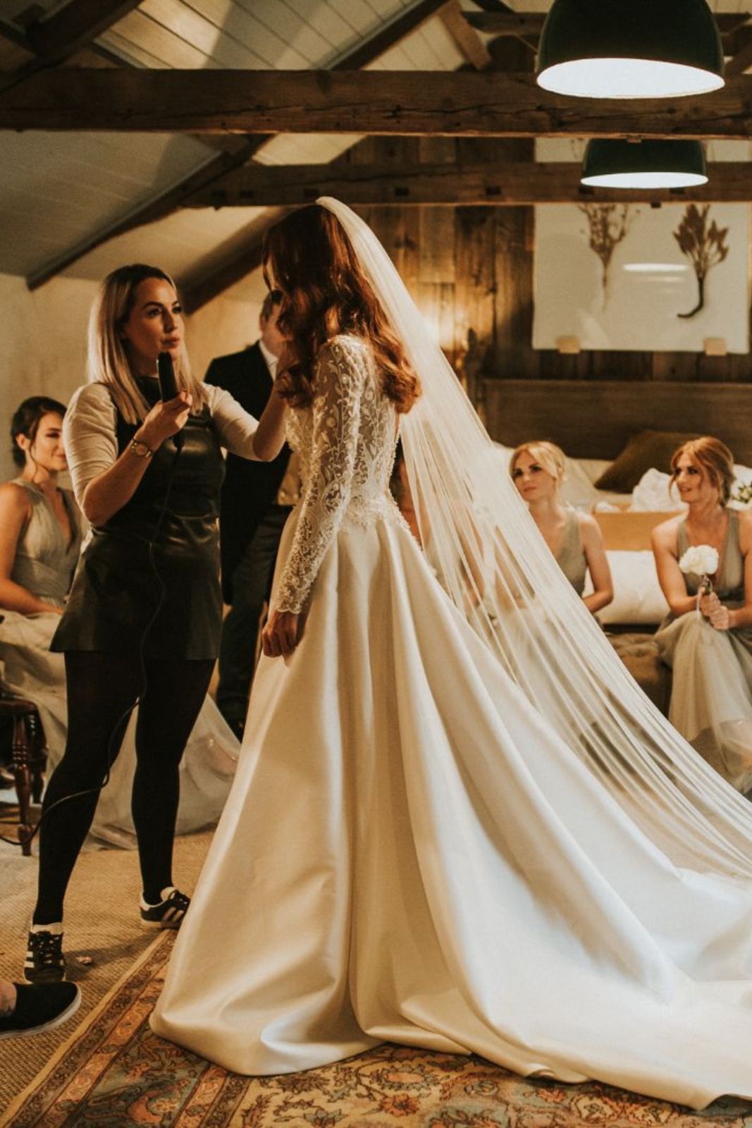 Blonde make-up artist touching up the Bride's face in a bedroom with wooden beams, bridesmaids on bed, taken by wedding photographer