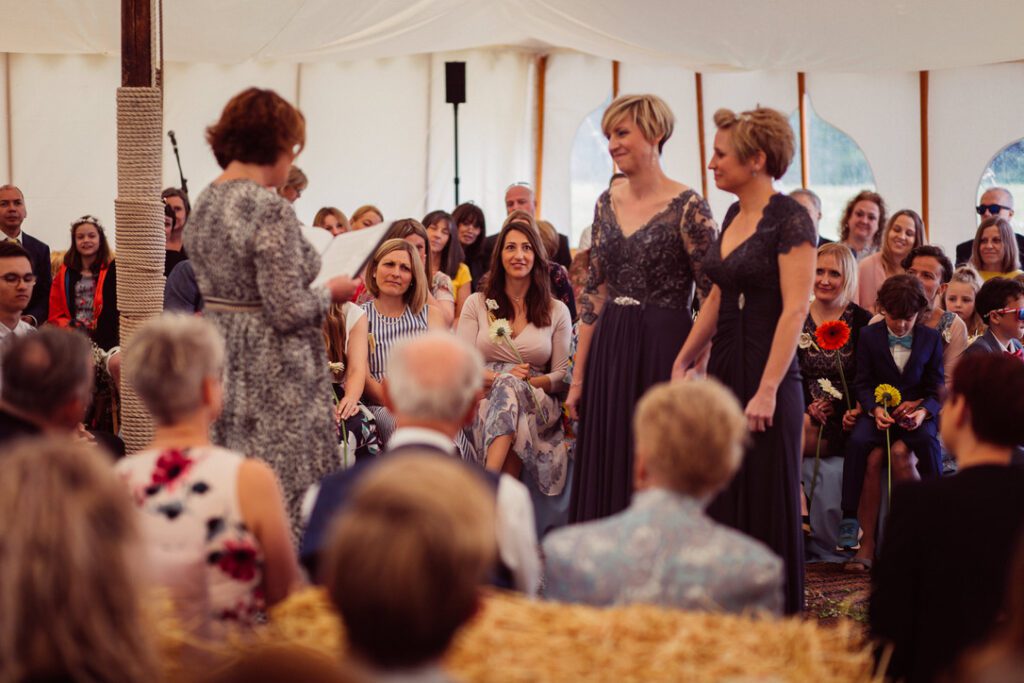 2 brides dressed in purple listening to fanfare ceremonies reading their wedding ceremony script surrounded by their guests  seated on straw bales enjoying the content 