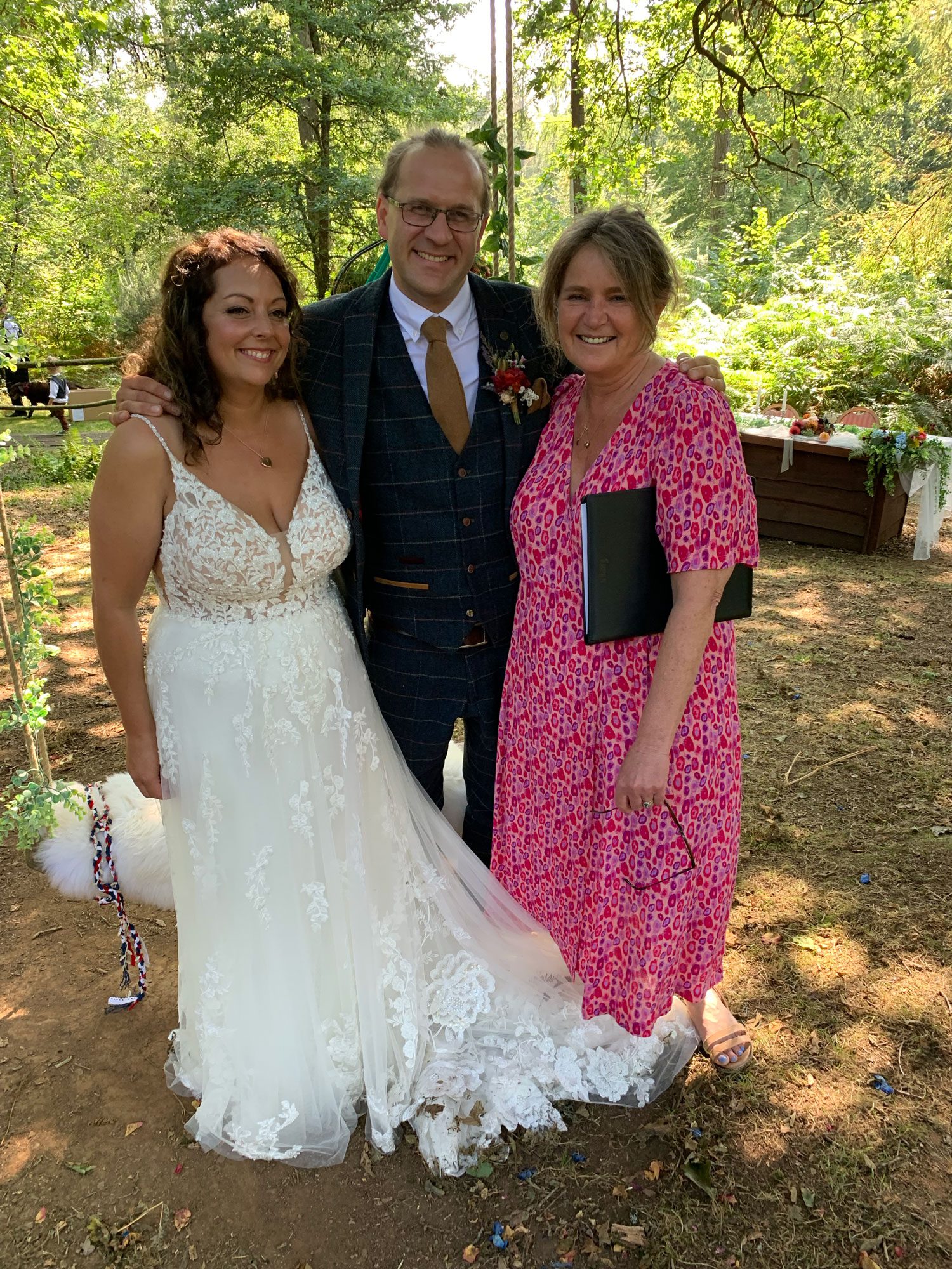 What does a wedding celebrant do? Well here, Fan in a deep pink floral dress is standing with her folder tucked under her left arm. To her right is the Groom in a dark checked suit and tan tie. To his Right is his bride in a strappy wedding dress with a deep v neckline. The top is covered in lace flowers, these are continued into the chiffon style skirt. They are posing under the canopy of trees on a sunny day.