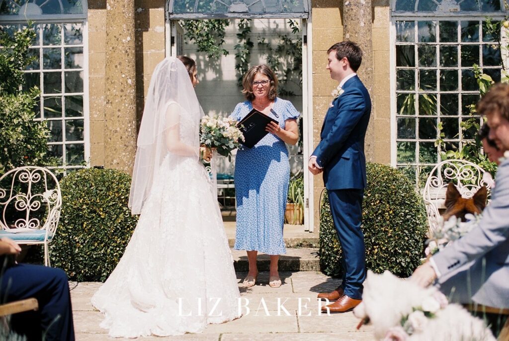 bride and groom standing looking at one another bride in beautiful white dress with a veil, groom in a suit celebrant Fan wearing a blue dressing all standing in front of an orangery