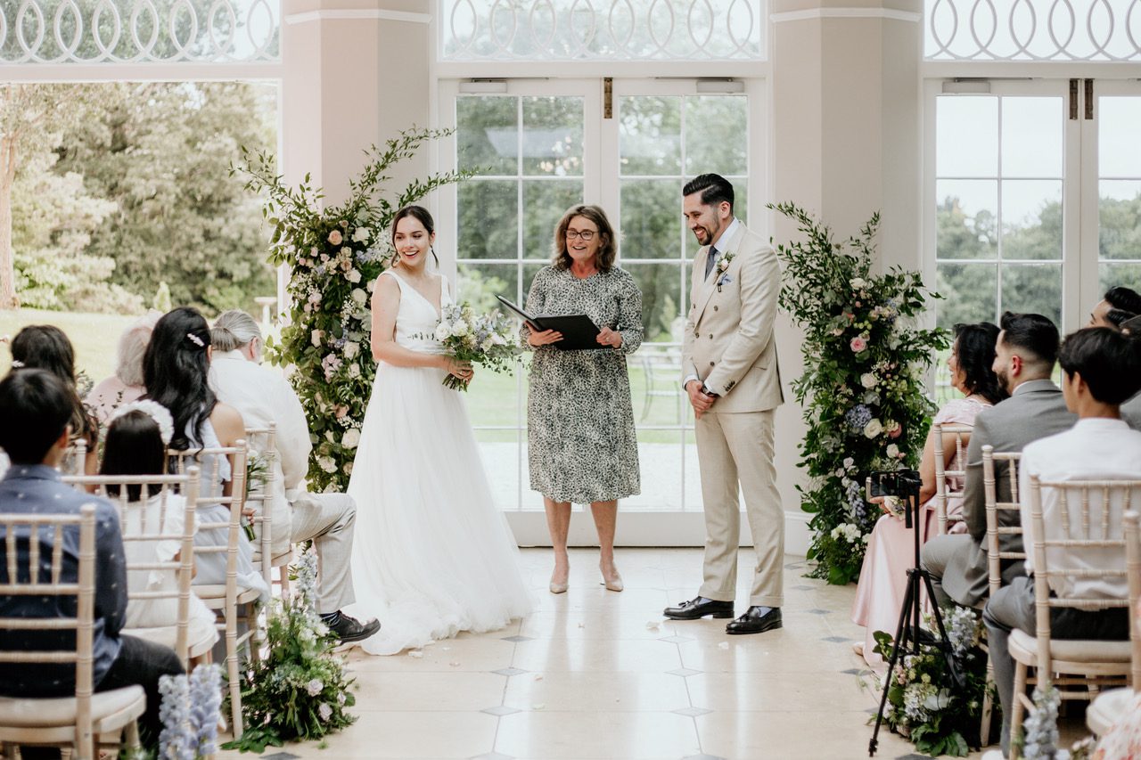 Friends are not great wedding celebrants, which is why in this image professional celebrant Fan is conducting the service which is being held in a brightly lit conservatory with beautiful cream flowers. The bride and groom are facing their guests laughing. The bride is wearing a long, simple, sleeveless gown with a crystal belt, the groom is dressed in a cream suit.