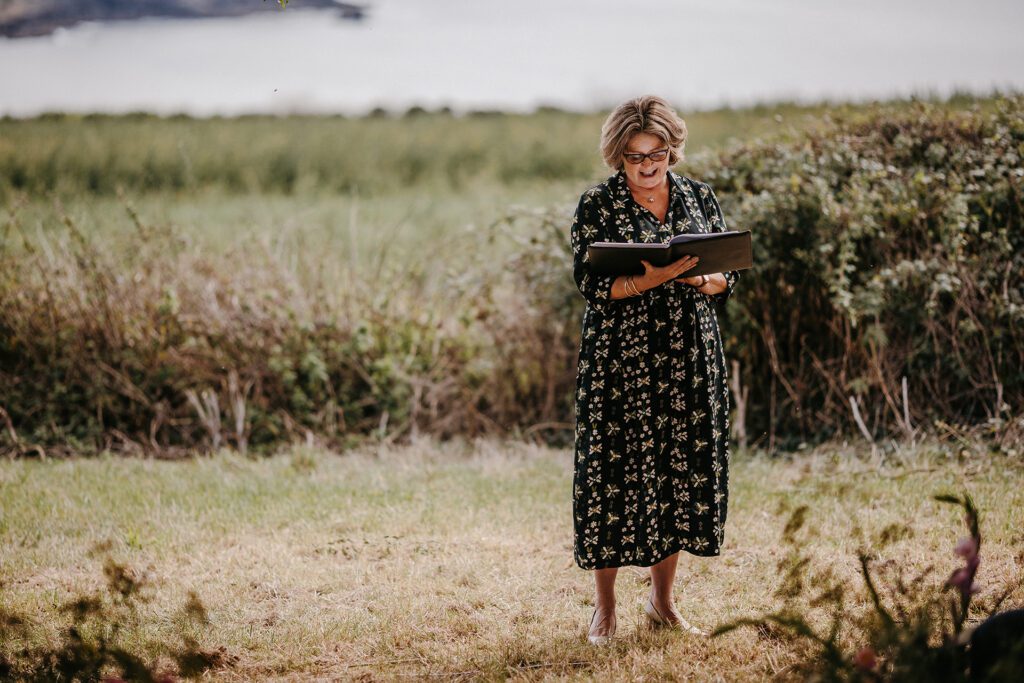 Wedding Celebrant Fan is stood in a field with hedges and more fields blurred in the background. Fan is holding an open black folder which she is looking down at and reading. Fan is wearing a long black dress with cream patination. Fan has short brown hair and is wearing glasses.