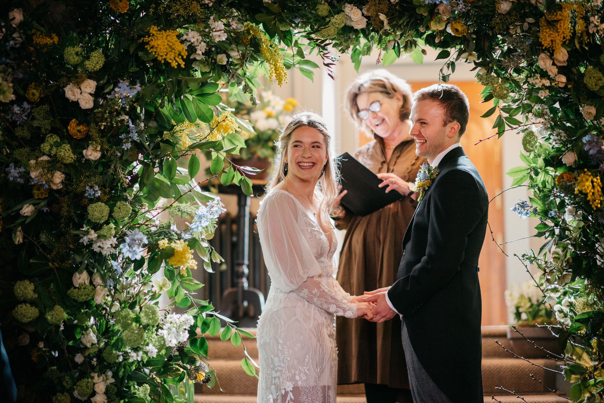 Personalised wedding ceremony by Fan, the celebrant, stood in the background in a copper coloured dress. The bride and groom are turned, facing their guests laughing. The groom is in a classic black morning coat, the bride is in a delicate lace fitted gown with transparent sleeves. They are under a beautiful golden, cream and green floral wedding arch