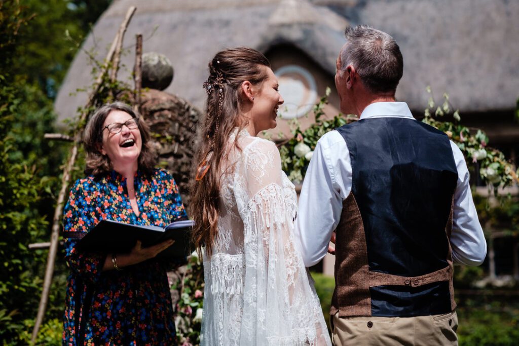 independent celebrants: Fan in blue floral dress laugh with bride in lace dress and groom in blue waistcoat at outdoor ceremony thatched building in background