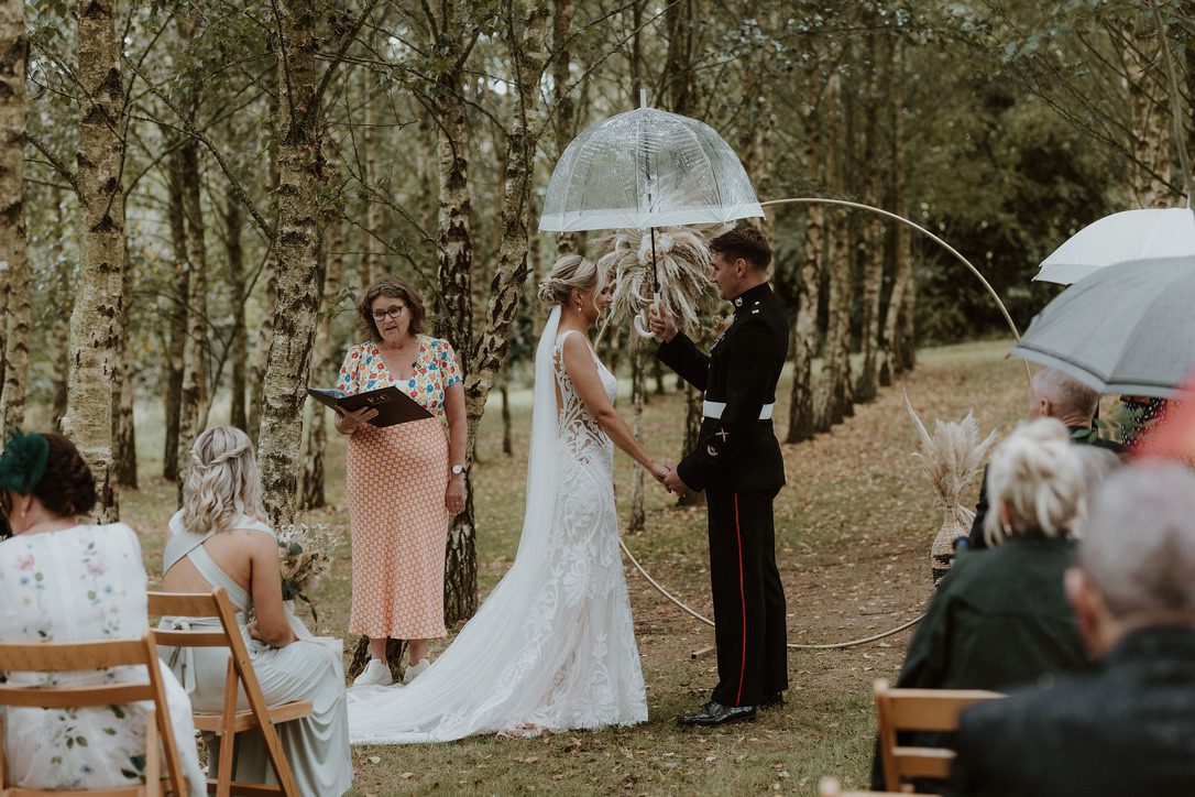 Fan, a wedding celebrant in Devon holding a ceremony for a bride and groom, stood under a clear umbrella as their guests watch