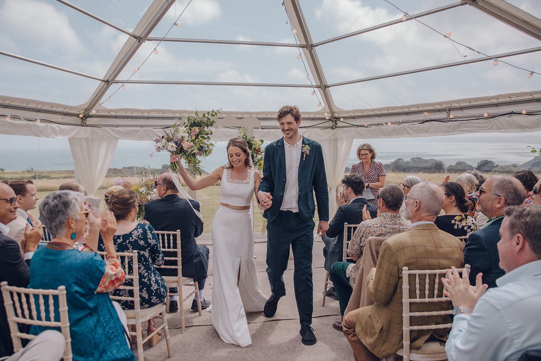 Your unique story  : Told by Celebrant Fan like this wedding under a marquee. The Bride in white fitted plain gown, Groom in Casual Navy suit are passing their seated guests.
