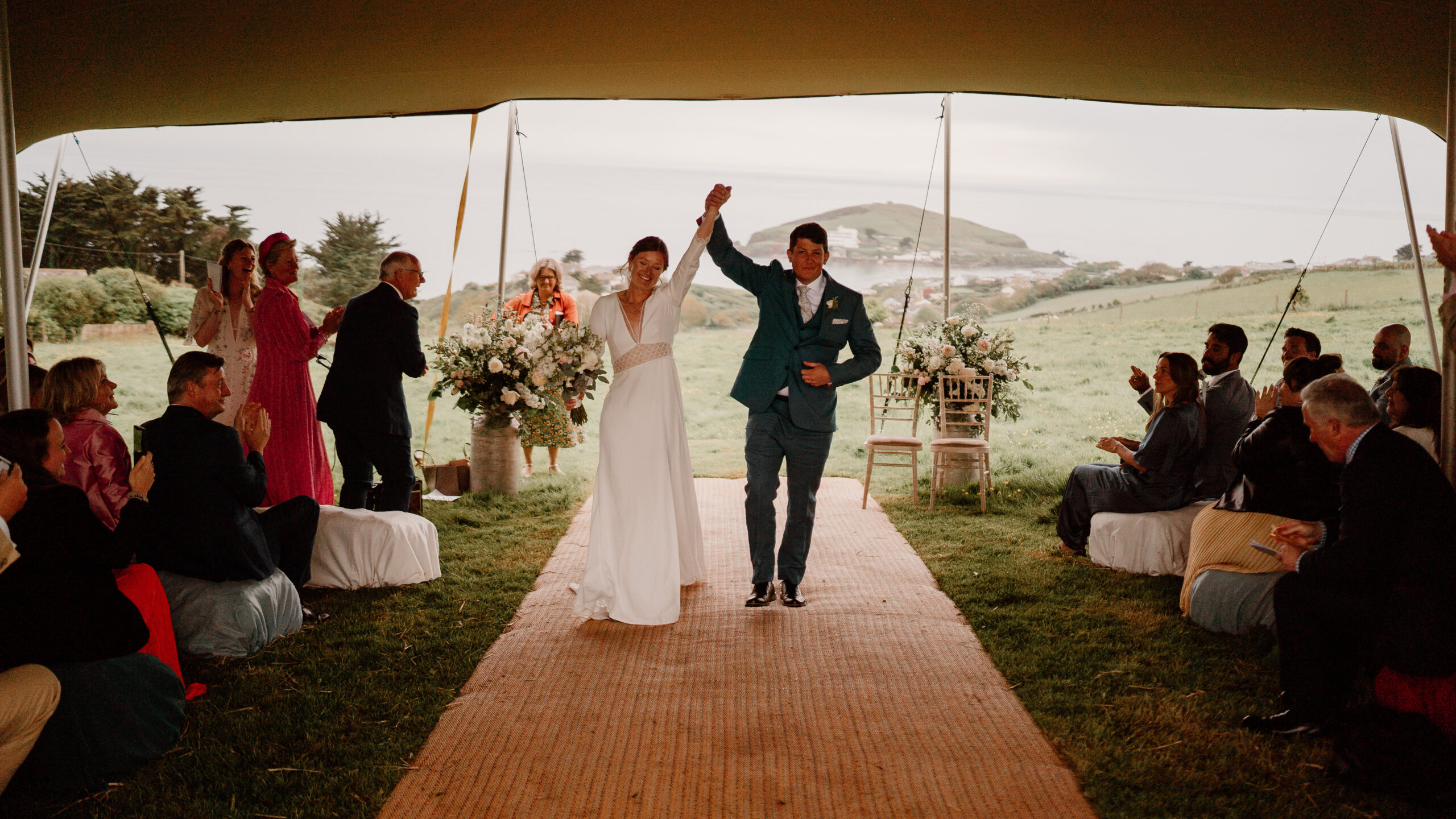Your unique story : a very happy couple leaving their wedding ceremony with their guests cheering, under a marquee, overlooking the sea.