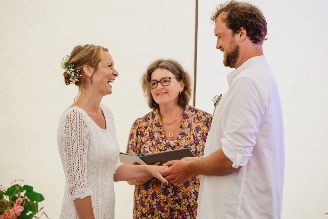 2D8529FC-D025-4A67-9778-43E2050D84DE_1_105_c Bride in simple white dress with groom who is placing ring on her finger celebrant Fan standing between reading from her book in a floral jumpsuit