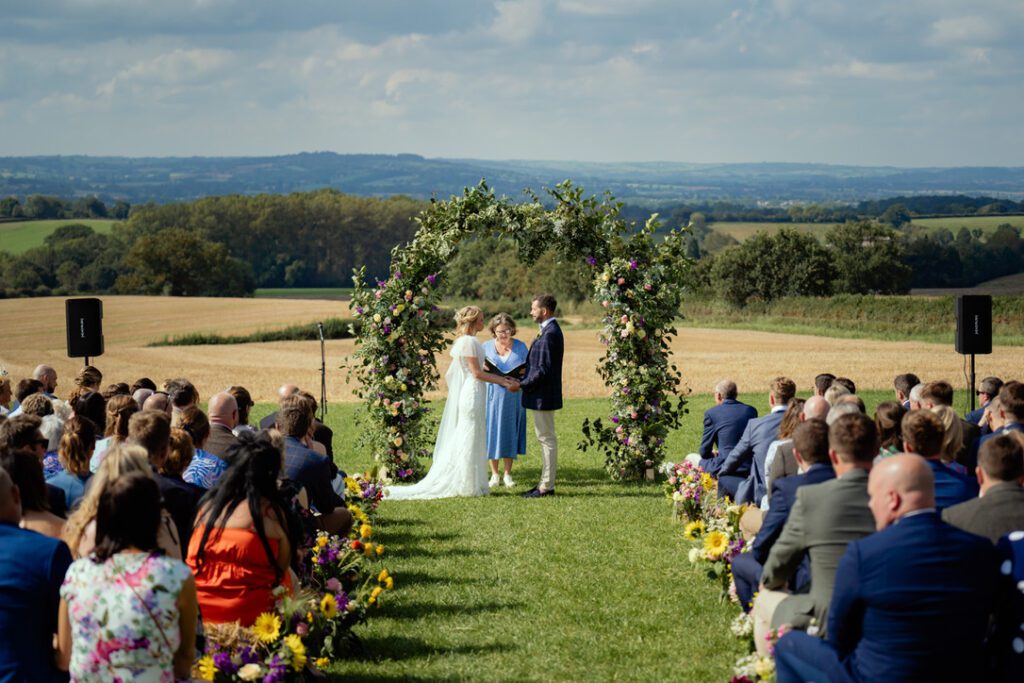Wedding at home - Louisa and Dan get married in field under floral arch, guests sit on bales of hay