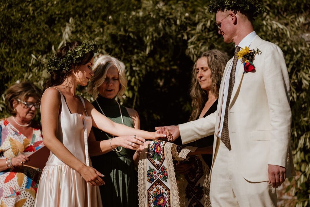 groom in white linen suit and bride in pretty white dress taking part in a hand fasting ceremony using a traditional ukrainian cloth with celebrant and relatives looking on 