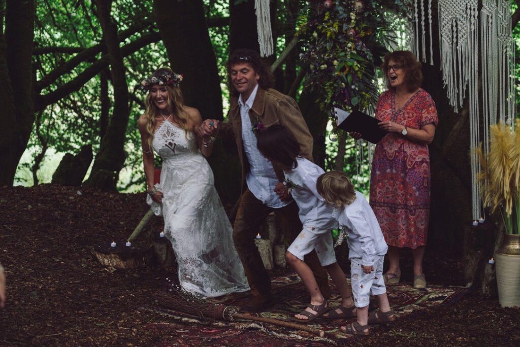 a couple and their two sons taking part in a jumping the broom ceremony in a woodland ceremony space with celebrant 