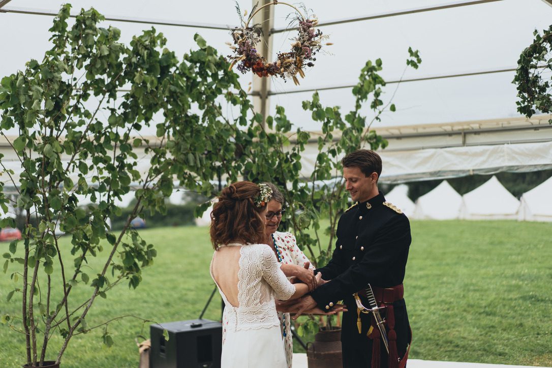 groom in uniform bride in white low backed dress pictured with celebrant who is placing their hands in correct position in preparation for a hand fasting ceremony