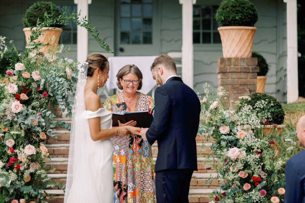 experienced wedding reading from her black book with bride with off the shoulder dress and groom standing at bottom of steps at Deer Park Country House