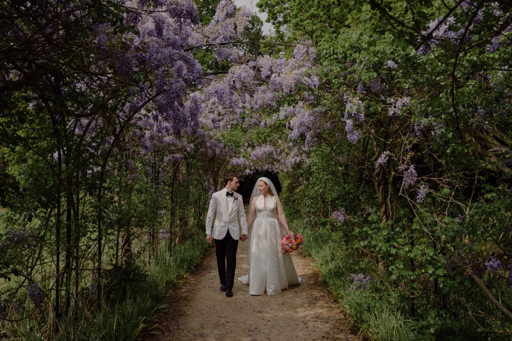 bride and groom walking under massive blue wisteria in full bloom in grove house gardens