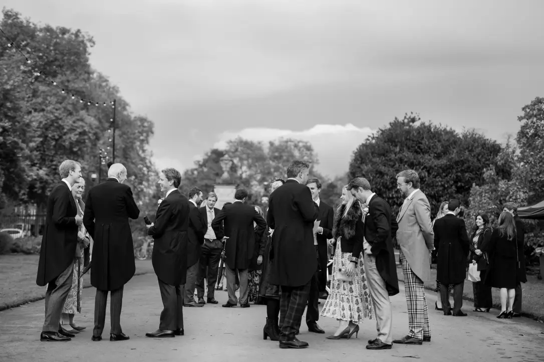 Black and white of wedding guests (men in morning suits) chatting on the driveway to a manor